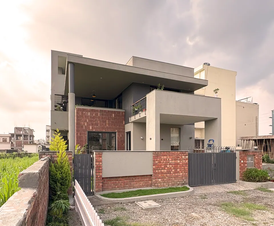 House with a deep cantilevered roof on a slender round column over a brick-clad block, behind a brick boundary wall and gate