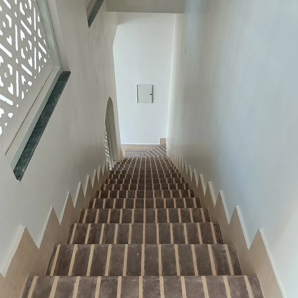 Narrow stair with dark grey stone treads and pale joint strips between white walls, lit through a jali screen window