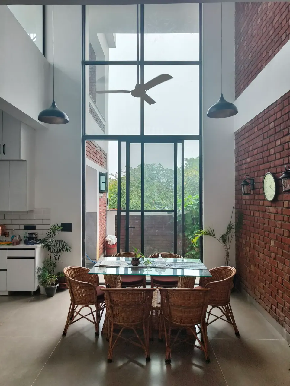 Double-height dining area with cane chairs around a glass table, full-height glazing, a brick wall and pendant lamps