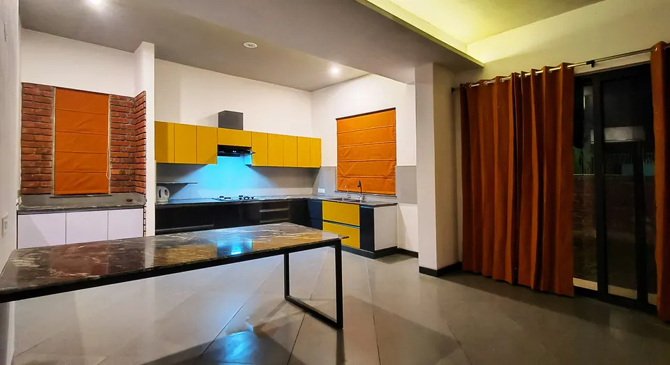 Kitchen with yellow wall cabinets, an orange blind on a brick wall, a long stone-topped table and orange curtains