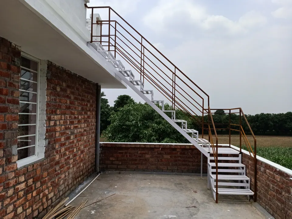 Roof terrace with brick parapets and a white steel stair with rust-red railings rising to the upper roof, fields beyond