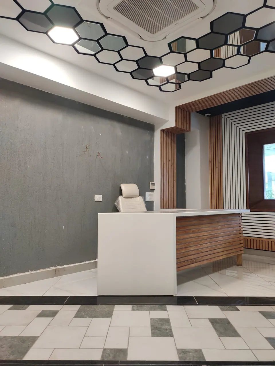 Reception counter with timber slats under a hexagonal mirrored ceiling, a grey textured wall and a patterned tile floor