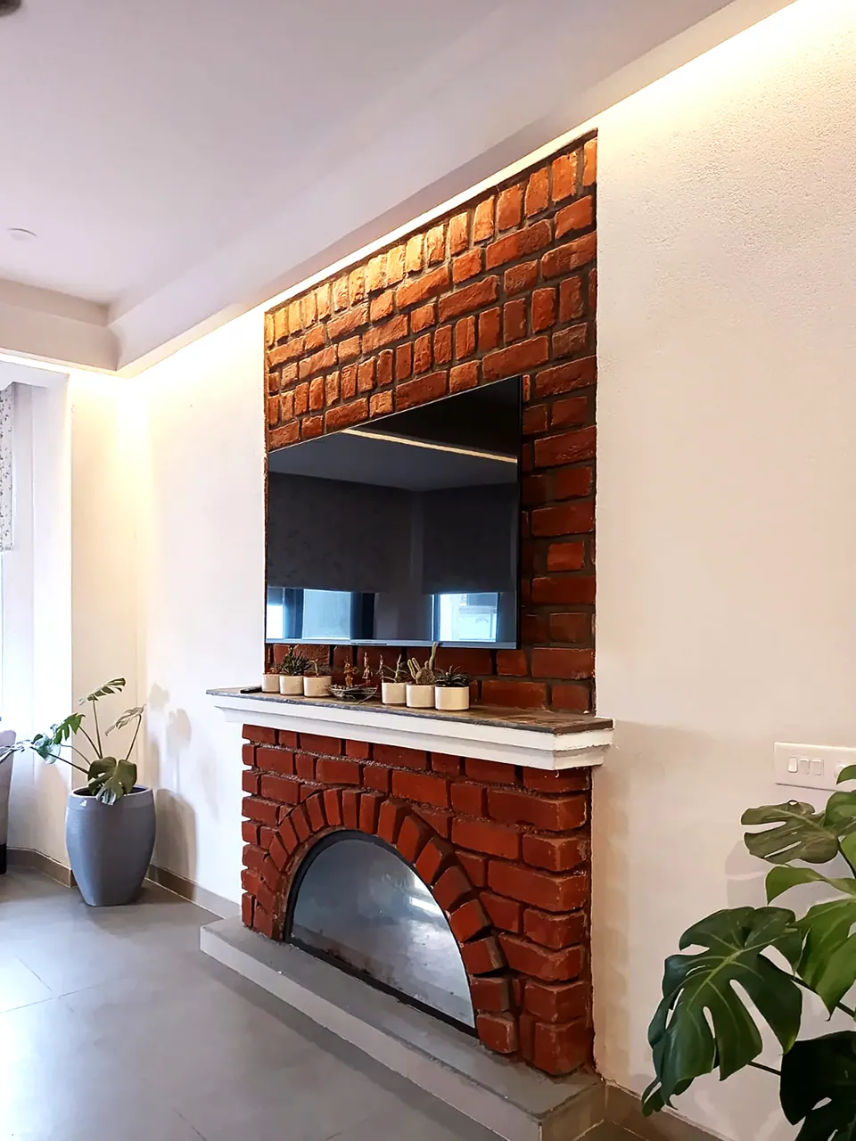 Brick chimney breast with an arched fireplace below a wall-mounted screen, potted plants alongside