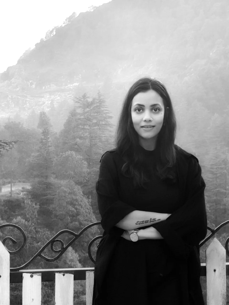 Black-and-white portrait of Gauri, arms folded, at a wrought-iron railing with a misty forested hillside behind