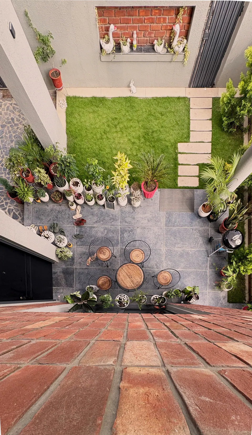 Top-down view from a brick-paved terrace into the courtyard: lawn, stepping stones, a patio table and potted plants